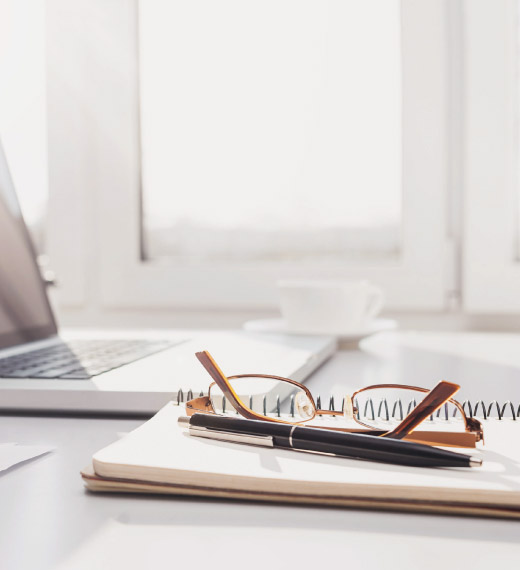 Computer, notepad, pen and eye glasses sit on top of a desk.