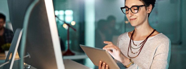 Woman wearing glasses sits in from of her computer holding a tablet.